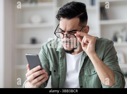 Problemi di vista concetto. Giovane uomo arabo in occhiali guardando lo schermo dello smartphone Foto Stock
