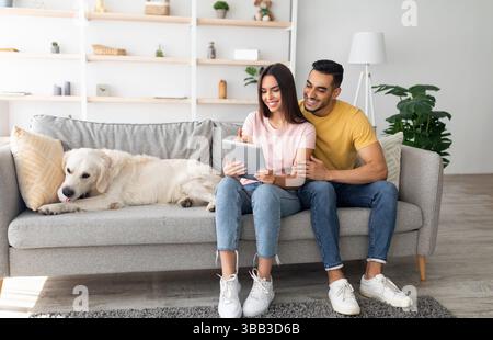 Bella giovane e variegata coppia seduta sul divano con il suo simpatico cane, che usa un tablet pc e naviga sul Web a casa Foto Stock