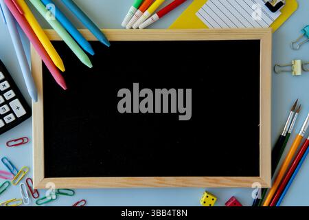Vista dall'alto della scrivania scolastica con lavagna penne vivaci clip per carta clip per rilegatura calcolatrice pennelli colorati dadi e appunti sulla superficie blu Foto Stock