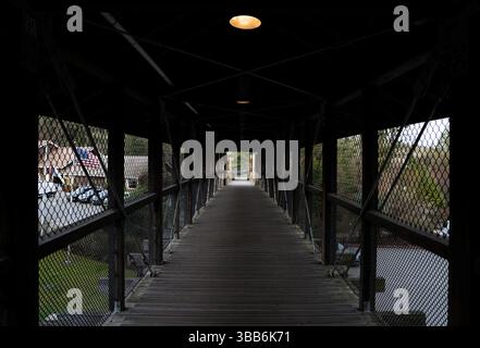 Una vista moody e simmetrica lungo un ponte pedonale coperto con maglie laterali e luci sopra la testa. Foto Stock
