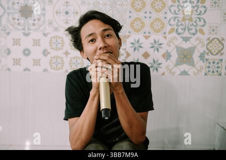 Uomo che canta con un microfono in mano, indossa una camicia casual, davanti a una parete a motivi vintage. Foto Stock