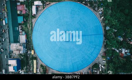 Uno scatto aereo di una massiccia struttura circolare blu vibrante, perfettamente centrata, circondata da vegetazione lussureggiante e vita urbana. Foto Stock