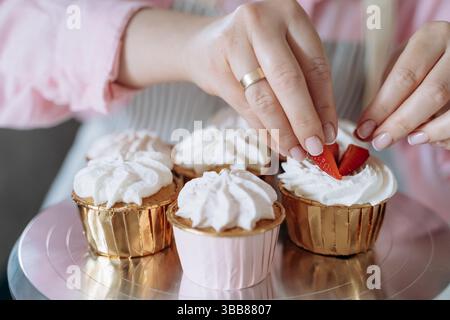 Prepara le mani fette di fragola fresche sui cupcake ricoperti di panna montata. L'immagine cattura il delicato processo di decorazione dei dessert per un vi Foto Stock