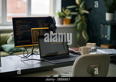 Area di lavoro moderna con configurazione a doppio monitor con codice di programmazione sugli schermi. Lo spazio di lavoro include laptop, monitor desktop, cuffie e altri uffici Foto Stock