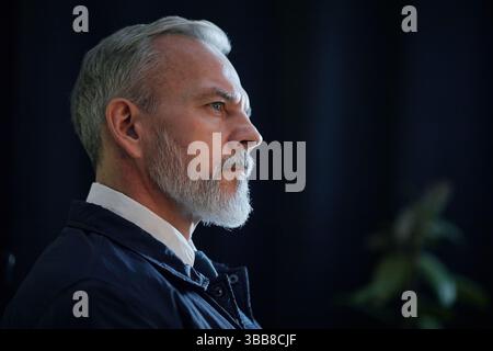 Uomo anziano con capelli grigi e barba che sembra pensivo mentre si siede su uno sfondo scuro. Luce che mette in evidenza il suo profilo e l'espressione contemplativa Foto Stock