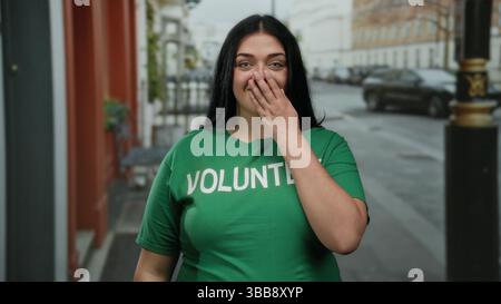 Donna sorridente che indossa una camicia verde volontaria in piedi per strada in città con una bocca che copre a mano l'energia giovanile e lo spirito della comunità all'aperto Foto Stock