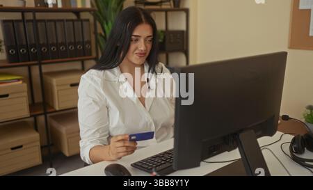 Donna che detiene la carta di credito in un ufficio moderno, lavora al computer, mostra l'ambiente aziendale con elementi organizzativi, un ufficio vivace Foto Stock