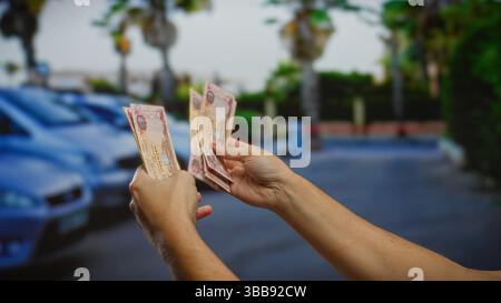 Uomo che tiene in mano banconote in dirham degli emirati arabi uniti sulla strada della città con auto parcheggiate nelle vicinanze che evidenziano le transazioni commerciali all'aperto Foto Stock