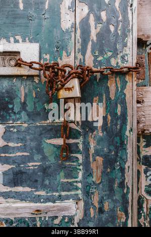 Portiera con agenti atmosferici fissata con catena Rusty e lucchetto. Primo piano di una porta in legno molto invecchiata con vernice blu e verde sbucciata, fissata a chiusura con un a. Foto Stock