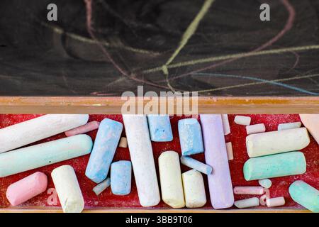 Pezzi colorati di gesso poggiano su un vassoio rosso sotto una lavagna, mostrando disegni di gesso in morbide tonalità pastello. Foto Stock