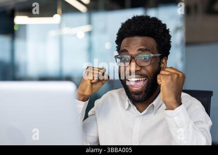 Un uomo afroamericano felicissimo celebra una vittoria guardando lo schermo del suo notebook in un ambiente d'ufficio. Il successo e la felicità si riflettono sul suo volto. Foto Stock