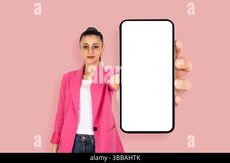 bruna ragazza che mostra un'applicazione mobile perfetta. Con uno smartphone moderno e di grandi dimensioni con un modello a schermo vuoto. Indossa una giacca rosa, con l'aspetto di una fotocamera. Foto Stock