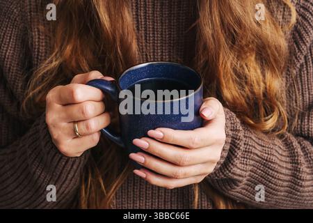 Donna che tiene la tazza da caffè. Bevanda calda in tazza blu. Maglione confortevole e capelli lunghi. Rilassante bevanda mattutina. Mani femminili con manicure. Foto Stock