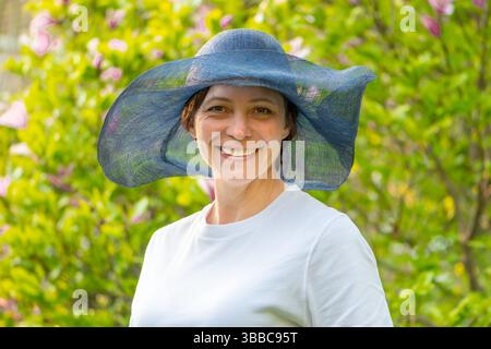 Una donna felice che indossa un ampio cappello blu posa di fronte a un giardino fiorito in una giornata di sole Foto Stock
