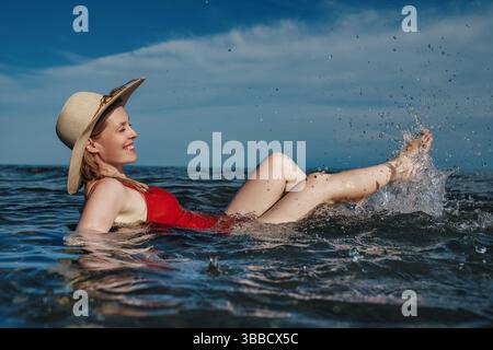 Giovane donna felice in costume da bagno rosso e cappello che spruzza in mare Foto Stock