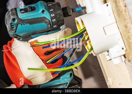 Un elettricista esperto utilizza un trapano elettrico per fissare i fili in una scatola di derivazione montata su una parete di legno. I fili elettrici sono colorati Foto Stock