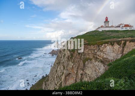 Una pittoresca vista costiera mostra un faro di Farol do Cabo da Roca arroccato su una verde scogliera che si affaccia su un mare turbolento. Le onde si schiantano contro Foto Stock