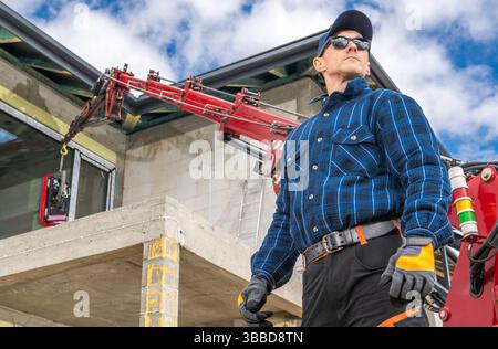 Un operaio edile attento si trova in loco con sicurezza, indossando una camicia a scacchi blu e un equipaggiamento di sicurezza. Guarda come una gru è in funzione, Lifti Foto Stock