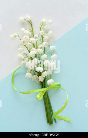 Primo piano di un delicato bouquet di giglio bianco dei fiori della valle legato da un nastro verde, posto su uno sfondo diagonale blu pastello e bianco. Mi Foto Stock
