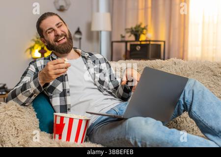 Giovane sdraiato sul divano di casa con ciotola di popcorn, ridendo mangiando i chicchi mentre guardi divertenti clip per laptop. Guy si scopa spontaneamente assaporando contenuti video stupidi che sprigionano un divertimento brillante. Foto Stock