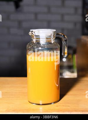 Un vaso di vetro trasparente pieno di succo d'arancia si trova su un tavolo di legno, catturato sotto una luce intensa in una cucina accogliente. Foto Stock