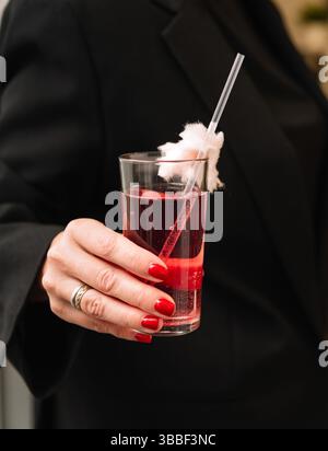 Elegante cocktail rosso frizzante servito in un bicchierino con zucchero di cotone sulla paglia, tenuto da una donna in nero. Foto Stock