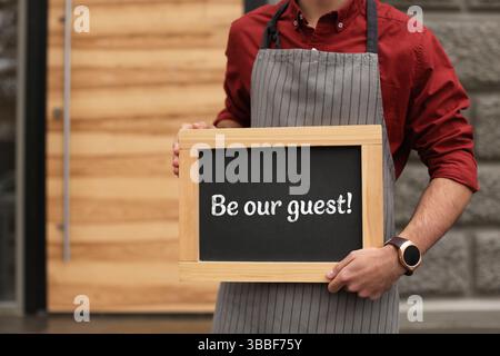 Uomo che tiene una piccola lavagna con la frase Be Our Guest Outdoors, primo piano Foto Stock