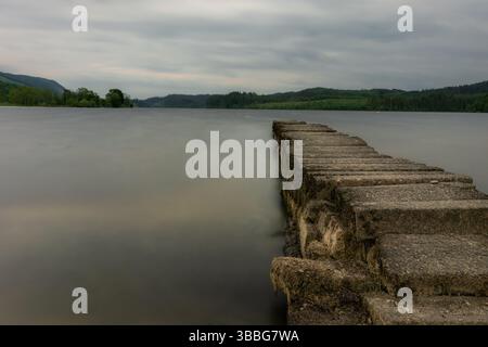 Un tranquillo molo si estende nelle nebbiose acque del Loch Ard, circondato da una foresta lussureggiante e da colline lontane sotto un cielo lunare. Foto Stock