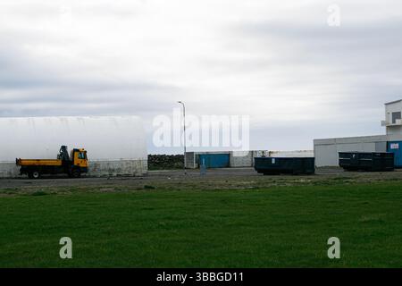 Akranes, Islanda - 15 maggio 2025: Una scena industriale rurale con un camion giallo, container e un magazzino bianco Foto Stock