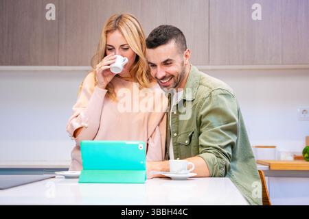 Coppia felice che usa il tablet insieme in cucina mentre beve caffè e sorride Foto Stock