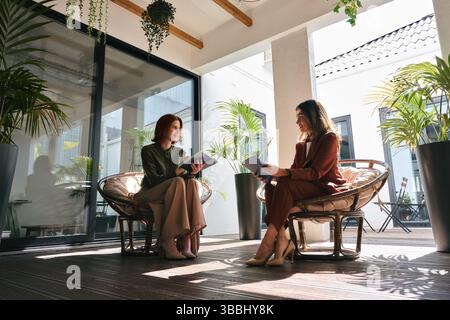 Due donne d'affari impegnate che hanno conversazione lavorando in un ufficio accogliente e verde. Foto Stock