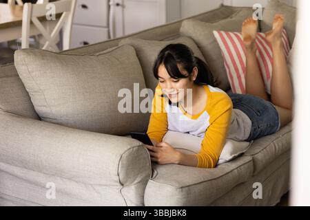 Donna asiatica che legge lo smartphone su un divano grigio chiaro nel soggiorno, con cuscino a righe rosa Foto Stock