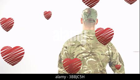 Soldato in piedi che indossa una giacca camo e un cappellino tattico sullo sfondo bianco, con disegni a forma di cuore rosso Foto Stock