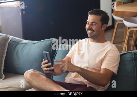 Uomo sorridente che tocca lo smartphone sul divano color tè nel soggiorno, con tavolo alto in legno Foto Stock