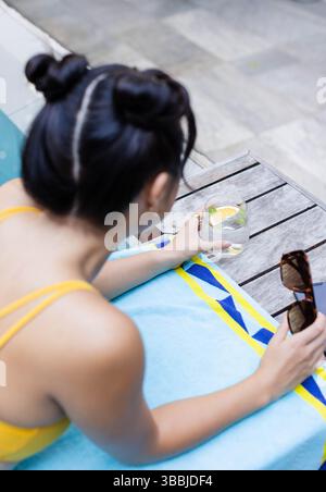 Posa di una donna asiatica in costume da bagno giallo appoggiata su un asciugamano sulla terrazza della piscina, tenendo acqua e occhiali da sole Foto Stock