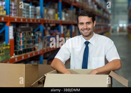 Uomo sorridente che maneggia scatole di cartone di grandi dimensioni nella corsia del magazzino, con scaffali in metallo Foto Stock
