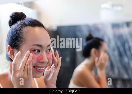 Applicazione donna asiatica che preme le toppe sotto gli occhi di idrogel rosa sullo specchio del bagno, copia spazio Foto Stock