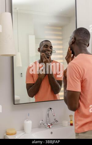 Sorridente uomo afroamericano di mezza età che tocca la guancia nel lavandino del bagno di casa, con specchio Foto Stock