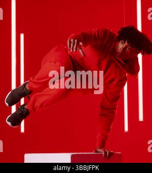 Ballerino afro-americano con cappuccio rosso in equilibrio sulla piattaforma in studio, con tubi LED Foto Stock