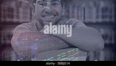 Uomo ispanico sorridente che indossa una camicia casual a maniche lunghe appoggiata al laptop in studio, con grafici delle scorte Foto Stock