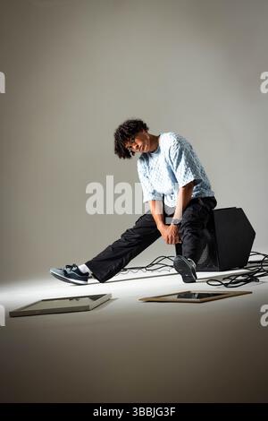 Uomo afroamericano pendente che si allunga le gambe sull'altoparlante in studio, con opere d'arte e cavi audio Foto Stock