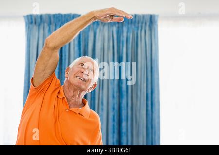Uomo anziano che indossa una polo arancione con braccio di sollevamento a casa con tende blu, spazio fotocopie Foto Stock