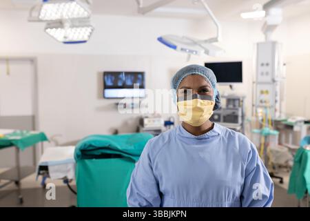 Infermiera donna multirazziale in piedi in sala operatoria, con lampade chirurgiche e apparecchiature sospese Foto Stock