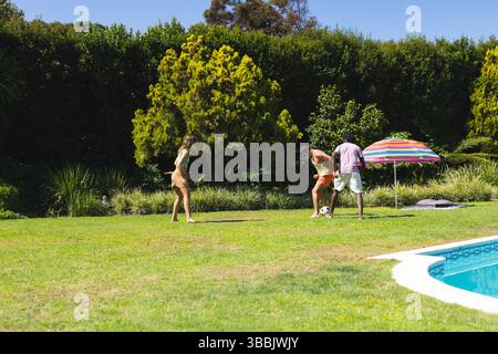 Giocare a diversi amici calciando palla da calcio sul prato vicino alla piscina, con ombrellone a righe Foto Stock