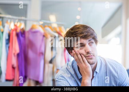 Un uomo pendente che tocca il mento in un negozio di abbigliamento, con camicie pastello su griglia metallica, spazio copia Foto Stock