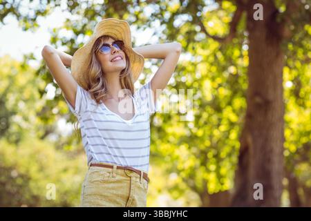 Donna che indossa un cappello di paglia e occhiali da sole sotto la luce del sole nel verde del parco con pantaloncini gialli Foto Stock