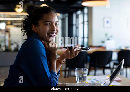 Giovane donna afroamericana adulta seduta a un tavolo in un ufficio aperto, con vivavoce vicino al computer portatile Foto Stock