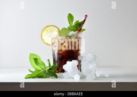 Bibita rinfrescante con ghiaccio in una tazza di vetro decorata con una paglia e una fetta di frutta su un tavolo bianco con frutta, ghiaccio e foglie con una iso bianca Foto Stock