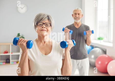 Allegra coppia senior che solleva manubri durante la riabilitazione del braccio in clinica, sorridendo e allenandosi insieme. Foto Stock