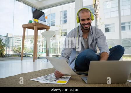 Uomo seduto che tiene in mano le foto stampate su un computer portatile in un ufficio moderno e indossa cuffie verdi al neon Foto Stock